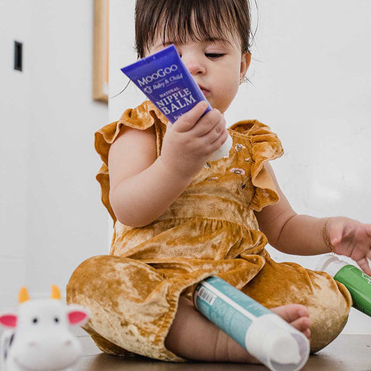 Child seated on floor holding MooGoo Baby & Child Natural Nipple Balm, lanolin free, gentle edible balm made with natural oils for breastfeeding mums