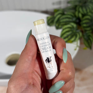 Hand holding a white tube of MooGoo Natural Coconut Cream Lip Balm with a plant and bathroom sink in the background.