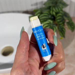 Hand holding a blue tube of MooGoo Natural SPF 15 Lip Balm with a plant and bathroom sink in the background.