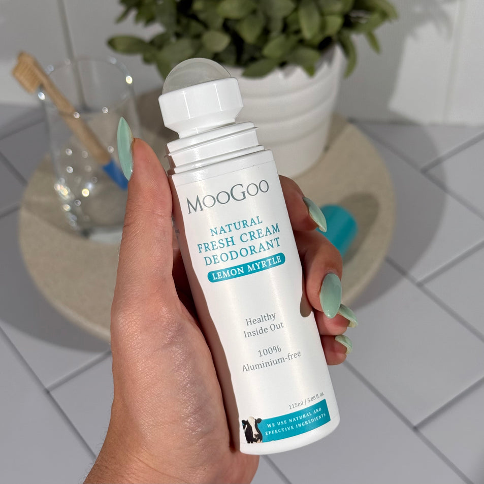 MooGoo Natural Lemon Myrtle Fresh Cream Deodorant 115ml held by a hand with a tiled background and a plant and toothbrush on a tray.