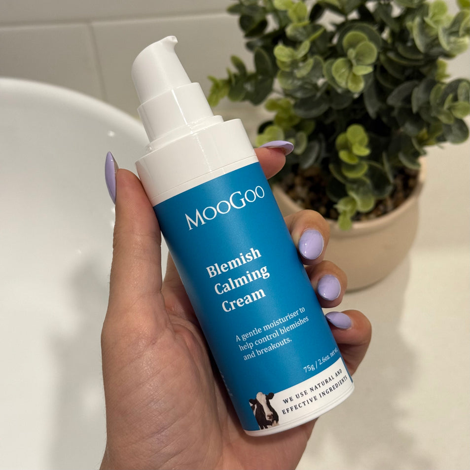 MooGoo Blemish Calming Cream in a blue bottle held by a hand with purple nails in a bathroom with a sink and plant in the background.