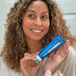 A smiling woman with curly brown and blonde hair holds a blue tube of MooGoo Peppermint Toothpaste in front of her face. The toothpaste packaging is blue with white text and a white stripe at the bottom.