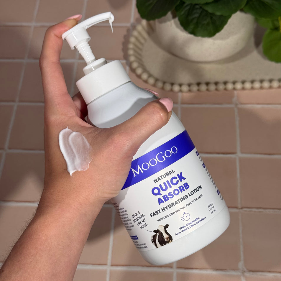 Hand holding a bottle of MooGoo Fast Hydrating Lotion with a swatch and a tiled bathroom background.