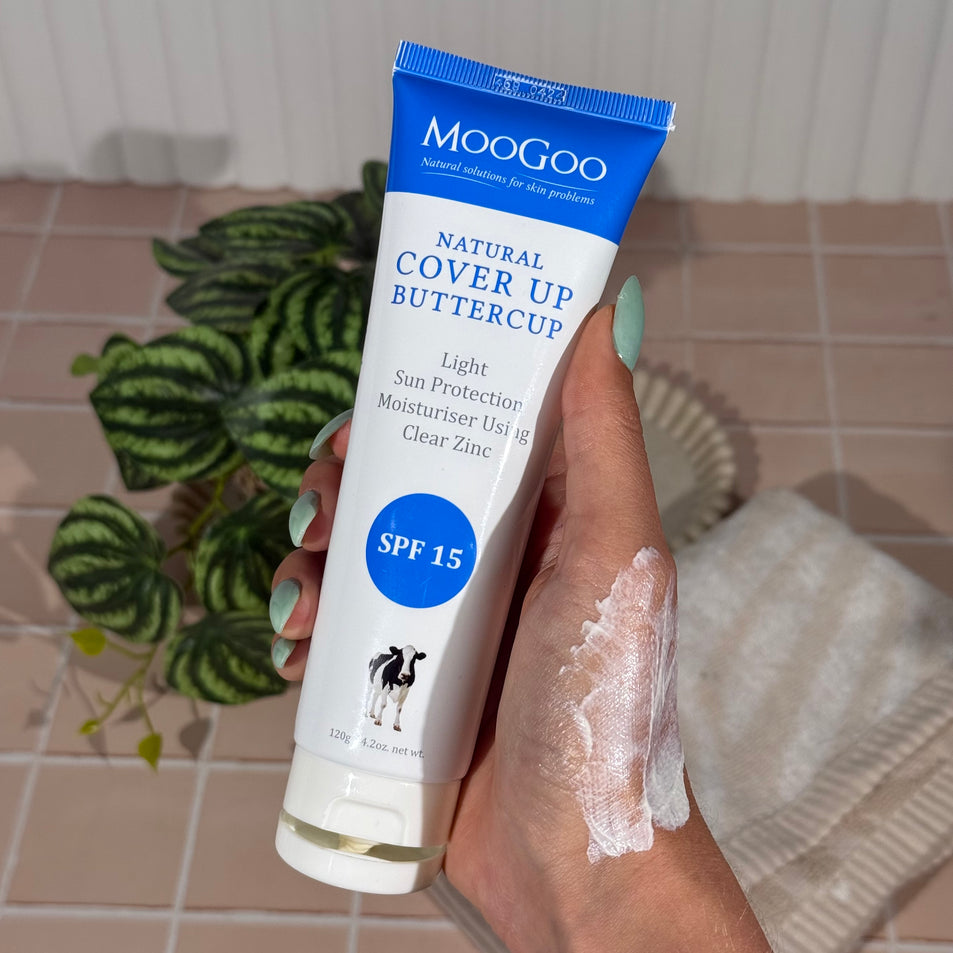Hand holding a tube of MooGoo Natural Cover Up Buttercup sunscreen with visible sunscreen on the hand, featuring a pink titled background with a plant, tray and towel.